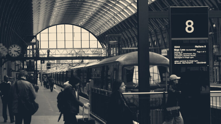 Commuters walk along a platform under King's Cross station's arched roof, past a train, clock and departure board.