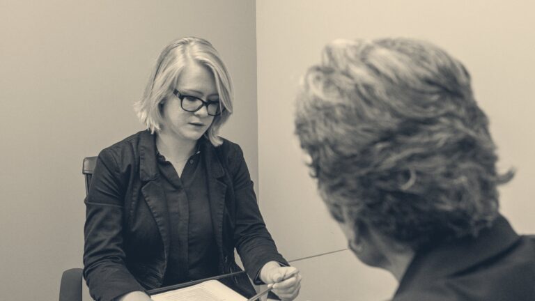 A woman in glasses and a black blazer reviews papers at a glass table, seen over the shoulder of the grey-haired hearing chair opposite her.