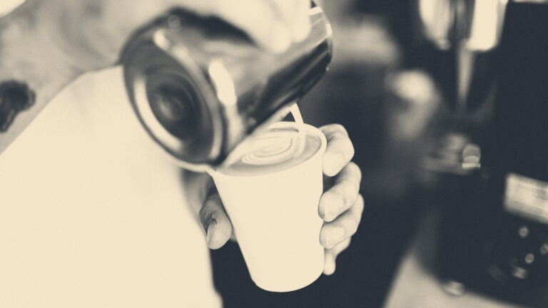 Barista in a white apron pours steamed milk into a takeaway cup, espresso machine behind.