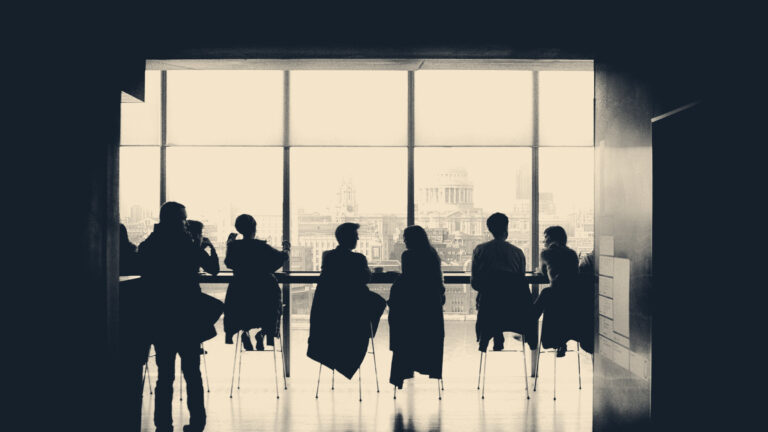Silhouetted colleagues sit at a long meeting table before a window overlooking the City of London.