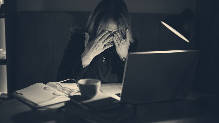 A woman sits at a desk at night, hands pressed to her face, behind an open laptop lit by a desk lamp