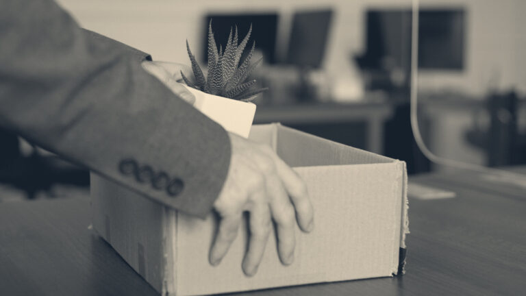 Hands in a grey suit sleeve packing a desk plant into a cardboard box, office desks behind.