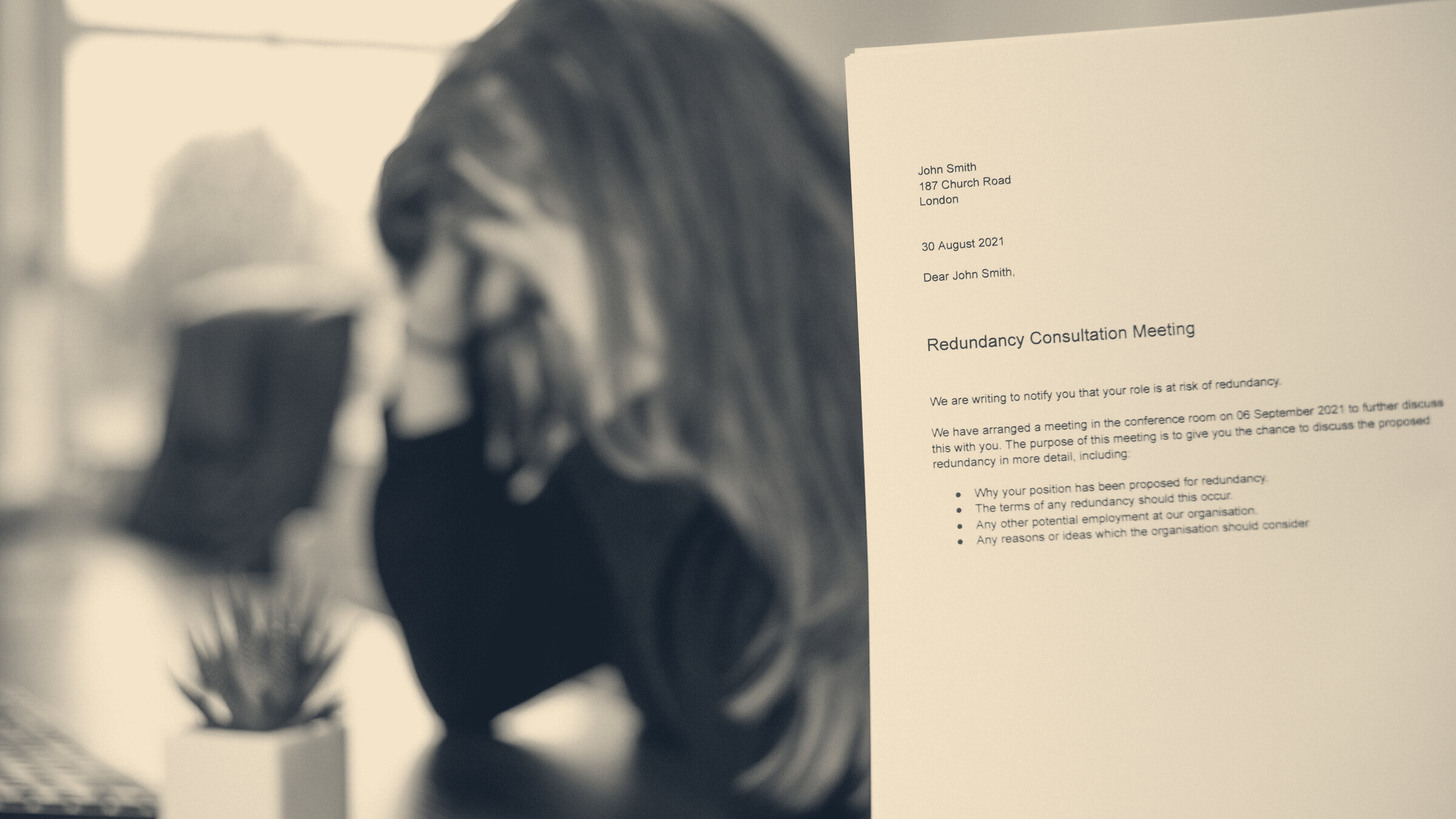 A dismissal letter titled 'Redundancy Consultation Meeting' in sharp focus beside a blurred worker with head in hand.