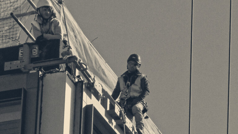 Two construction workers in harnesses, hi-vis and hard hats work high on the edge of a City of London building site.