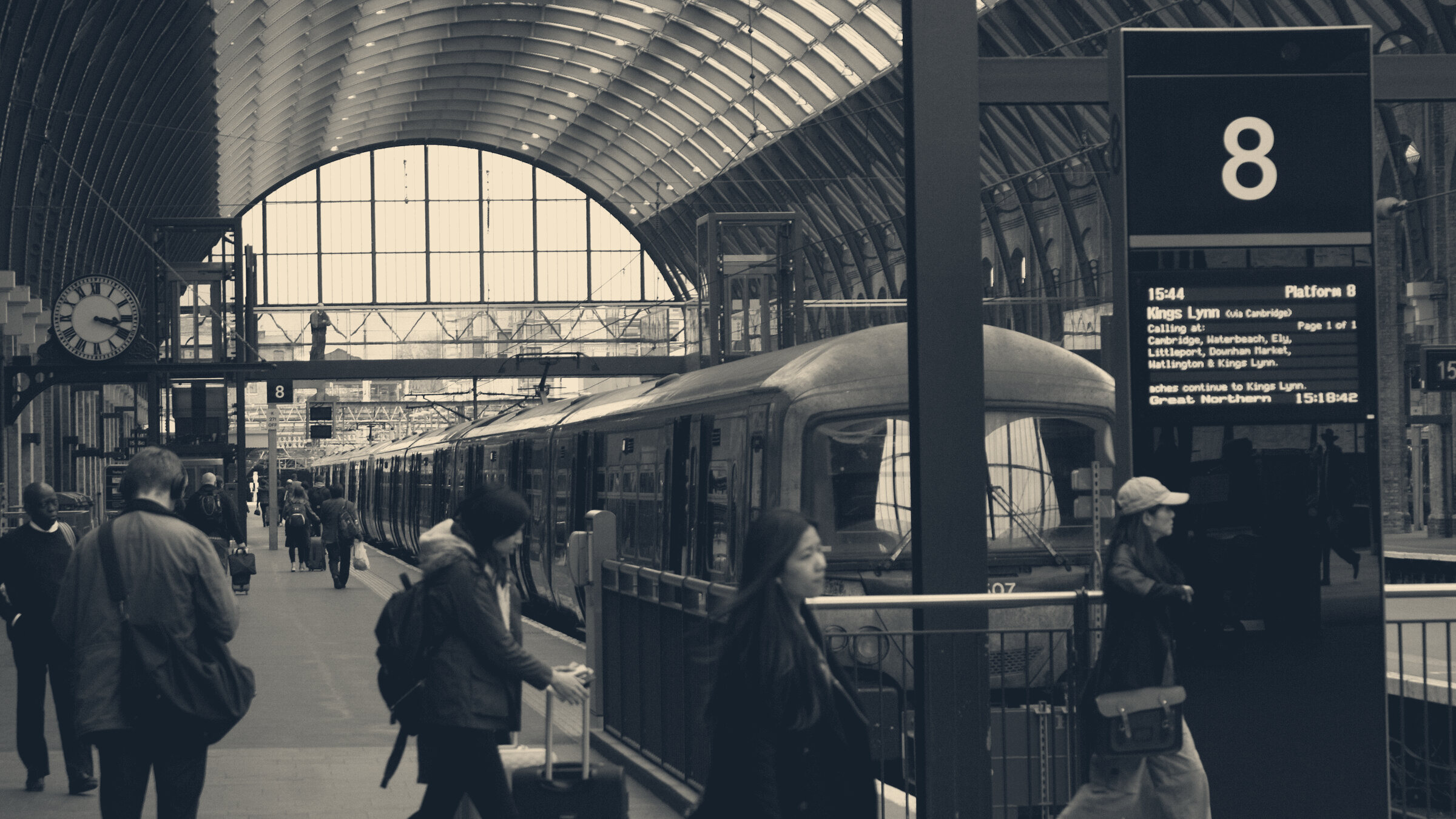 Commuters walk along a platform at London King's Cross station, beside a train and departure board.