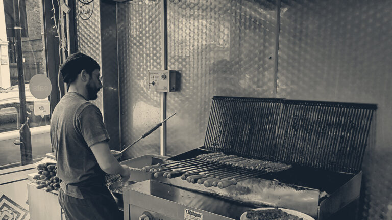 Cook in apron and cap grilling skewers at the counter of a London takeaway, street visible through the window.