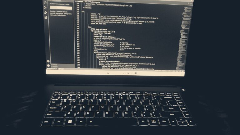 A backlit laptop keyboard glowing in a dark room, code on the screen — night-time computer work.