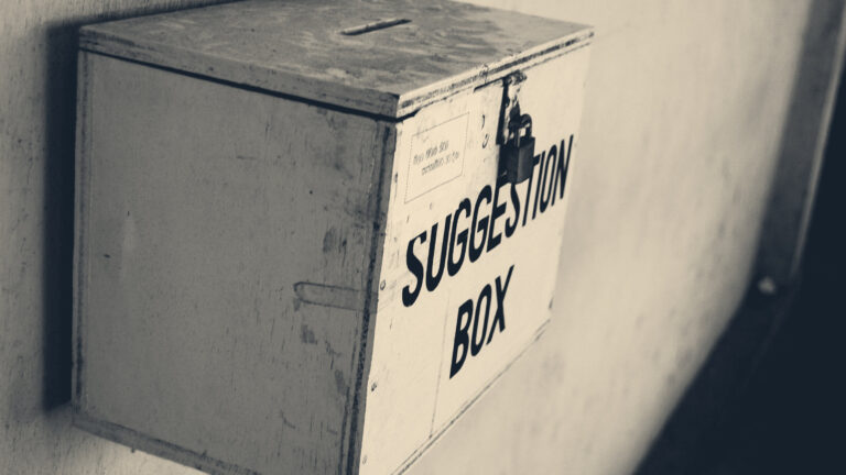 A dusty metal suggestion box with a brass padlock, mounted on a worn corridor wall.