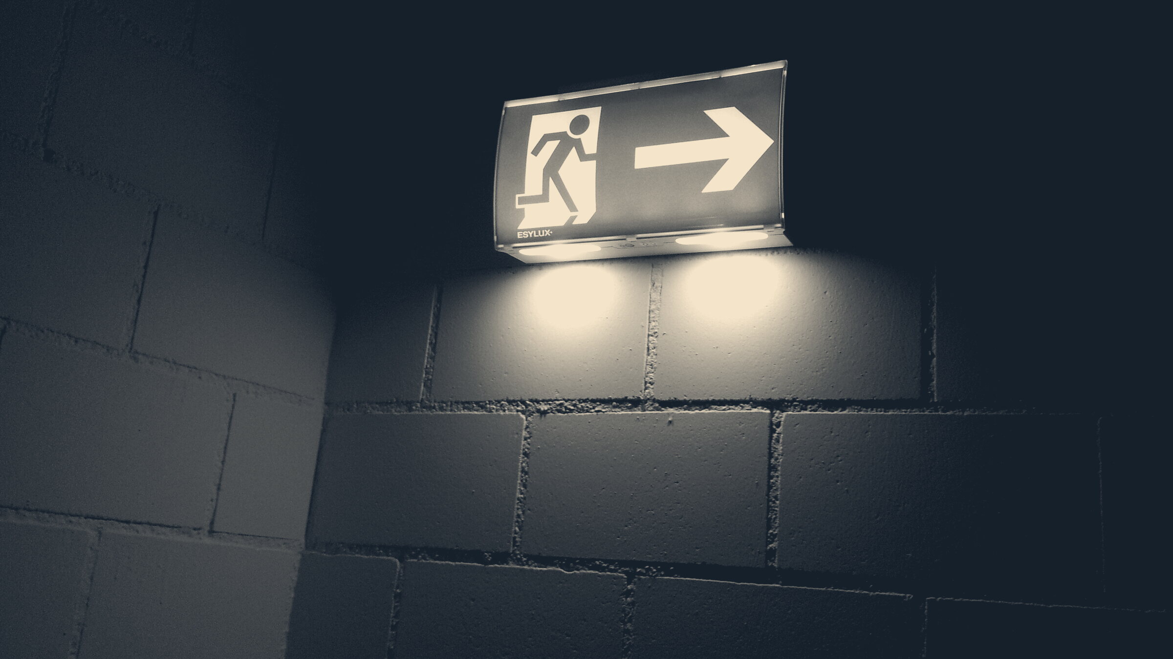 A lit green running-man emergency exit sign glowing on a painted concrete-block wall in a dark corridor.