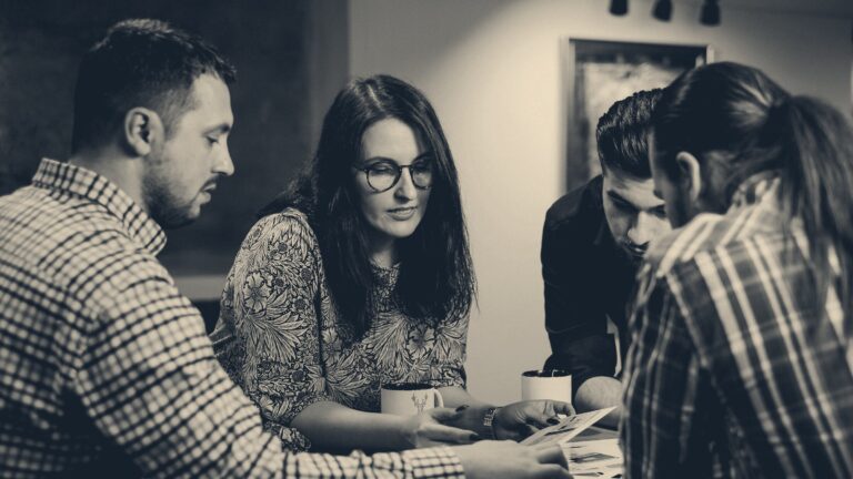 Colleagues lean over paperwork on a wooden table, one pointing at a document in discussion