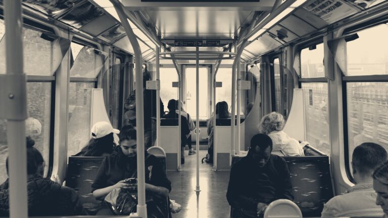 Commuters seated in a teal-railed London DLR train carriage, heads bowed over phones on the morning ride.