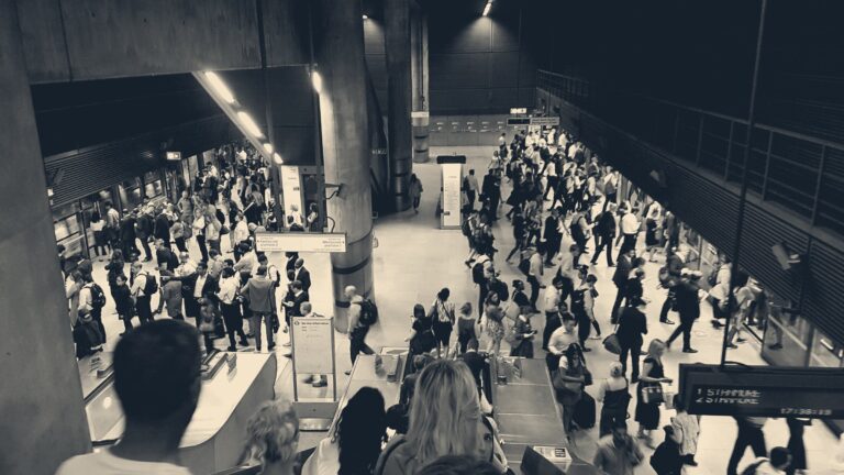 Commuters stream through the concrete hall of Canary Wharf station at rush hour, seen from the escalators above.