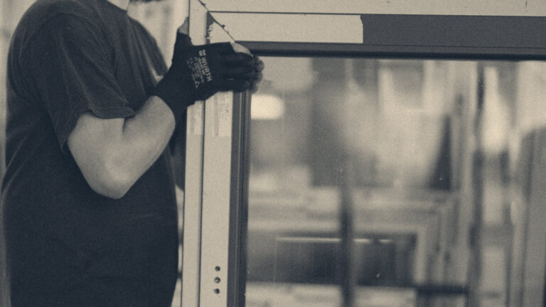 Gloved warehouse worker lifting a tall glass panel with both hands raised, dark workwear, blurred racks behind.