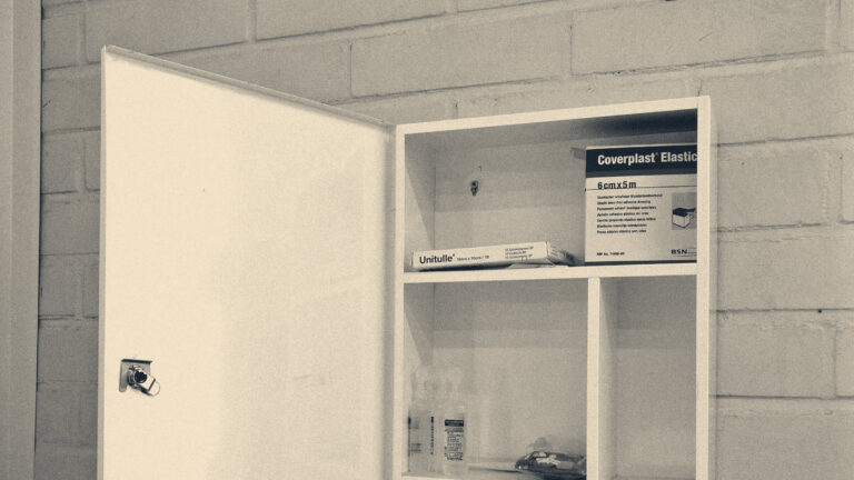 An open, nearly empty wall-mounted first aid cabinet holding a bandage box and dressing on a painted brick wall.