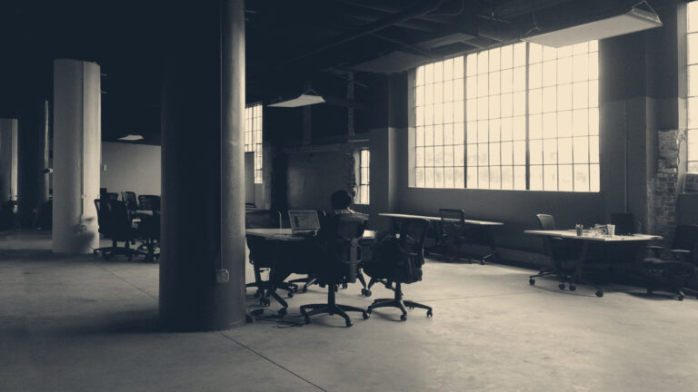 A lone worker in a hi-vis vest at a desk in a vast, dim industrial office beneath large grid windows.