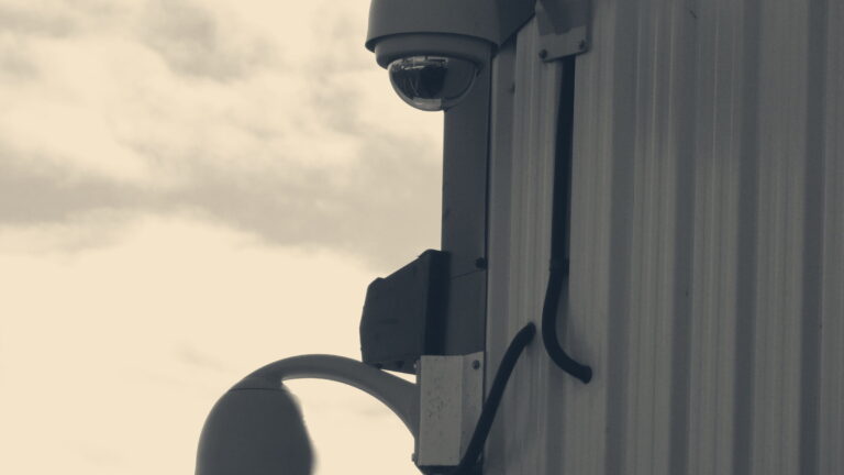 Two dome CCTV cameras mounted on corrugated cladding of an industrial building against a grey cloudy sky