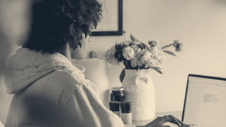 Over-shoulder view of a woman typing on a laptop at a home table beside a jug of flowers