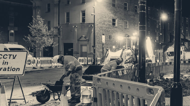 Two road workers in orange hi-vis shovelling behind blue barriers during night roadworks on a lit Kent street.