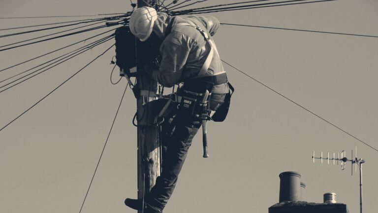 Telecoms engineer in high-vis jacket and safety harness working alone atop a wooden utility pole beside UK chimney pots.