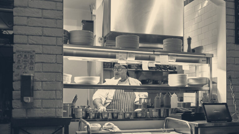 A chef in a striped apron works a stainless-steel pass hung with order tickets in a commercial kitchen.