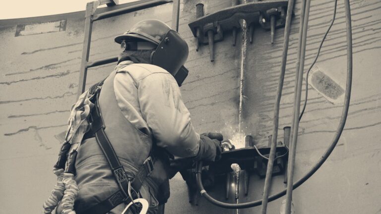 Welder in hard hat, face shield and safety harness welding a steel tank seam, sparks and fume rising from the arc