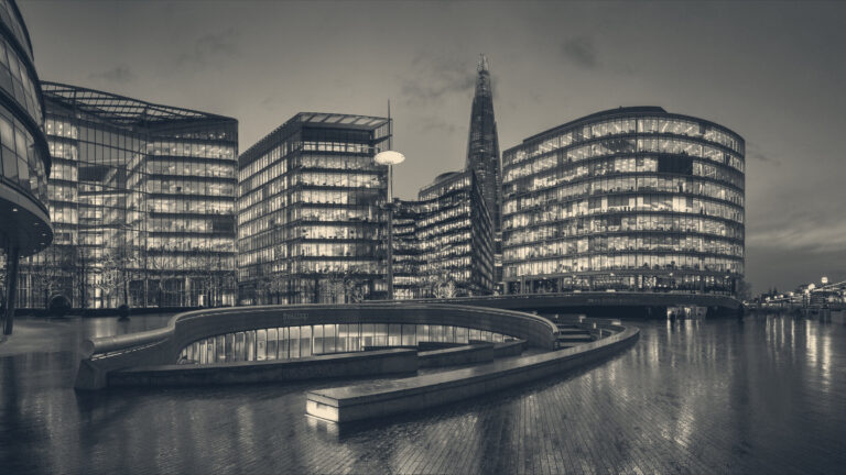 Lit office buildings at dusk along the Thames at More London, the Shard rising behind wet riverside paving.