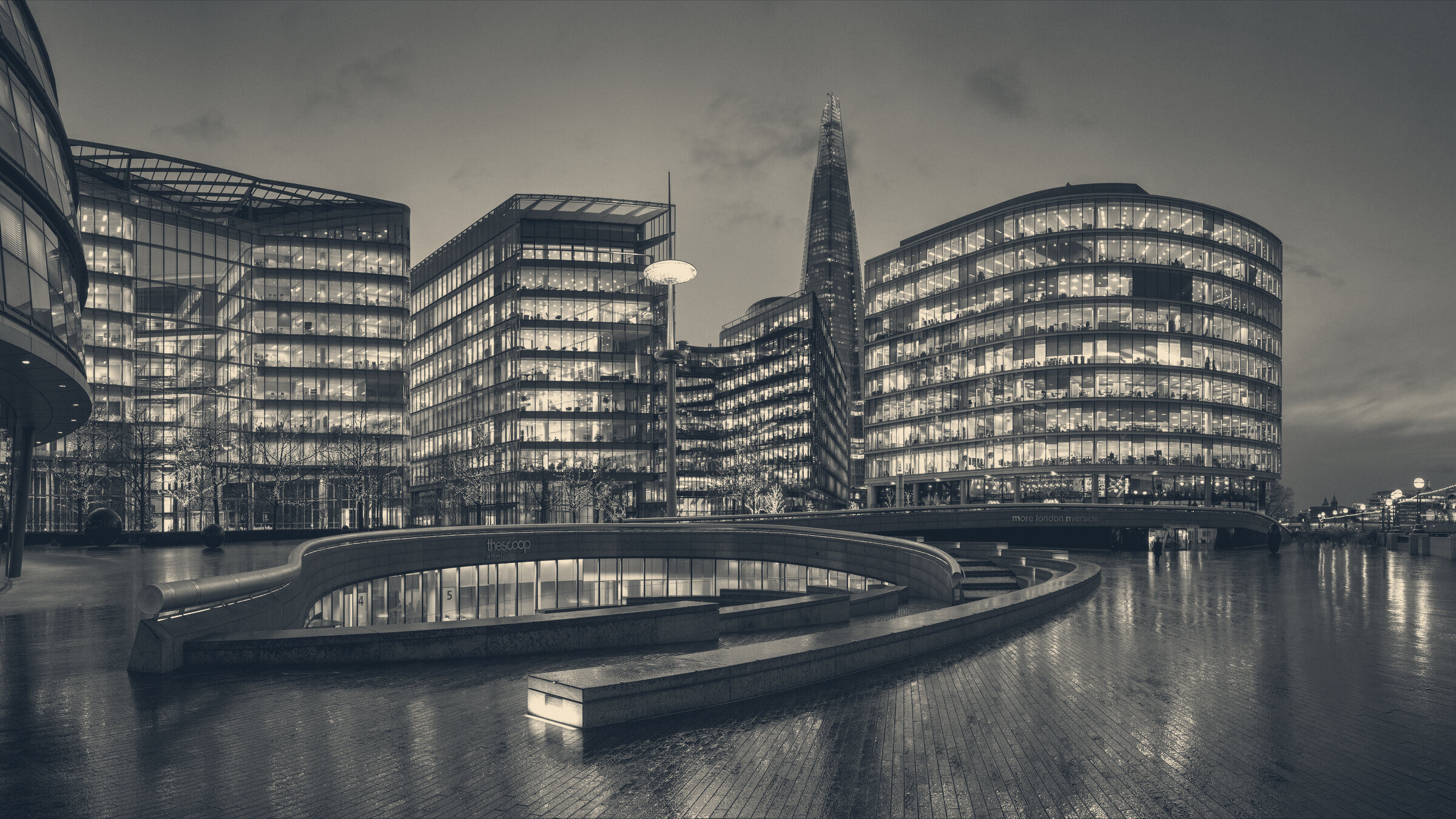 Lit office buildings at dusk along the Thames at More London, the Shard rising behind wet riverside paving.