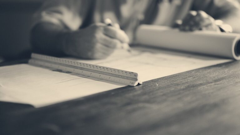 Architect sketching on blueprints at a wooden table with a triangular scale ruler