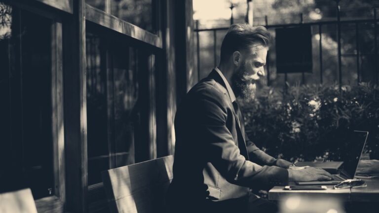 A man in a dark suit types on a laptop at an outdoor wooden cafe table in bright sunlight.