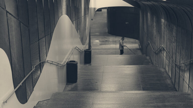 A lone maintenance worker in a yellow jacket mops at the end of a curved mesh-walled underpass lit by fluorescents.
