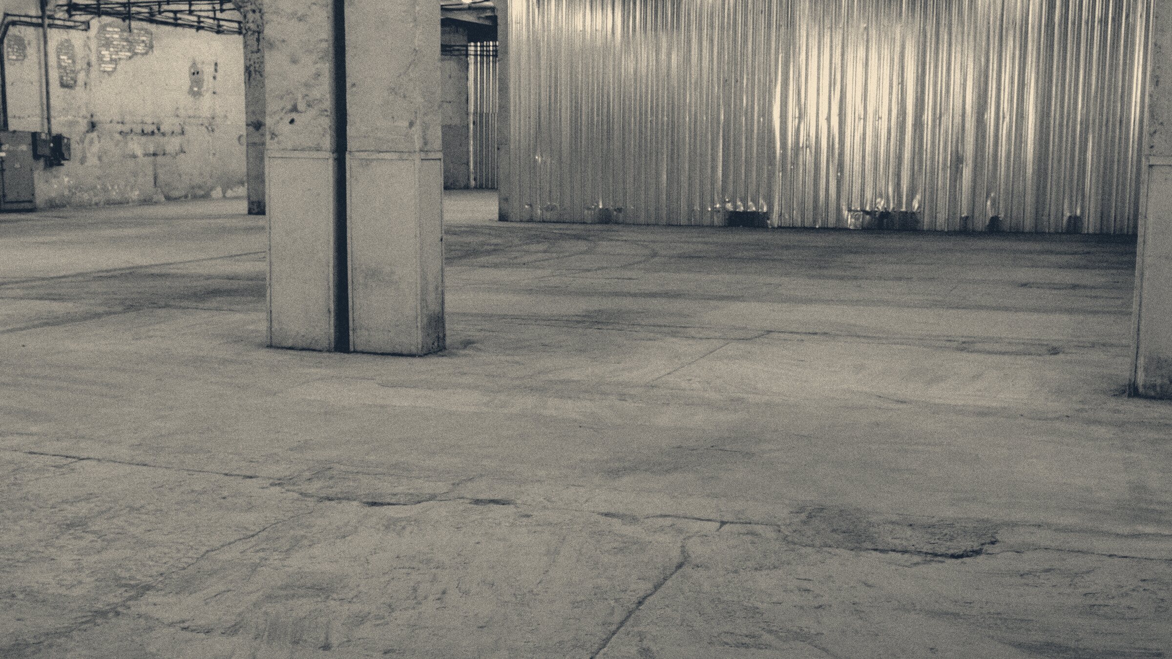 Empty warehouse interior: tyre-marked concrete floor, steel pillars and a corrugated metal wall.