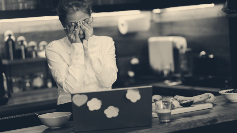 A woman at a kitchen table covers her face with both hands in front of a stickered laptop, coffee and fruit nearby.