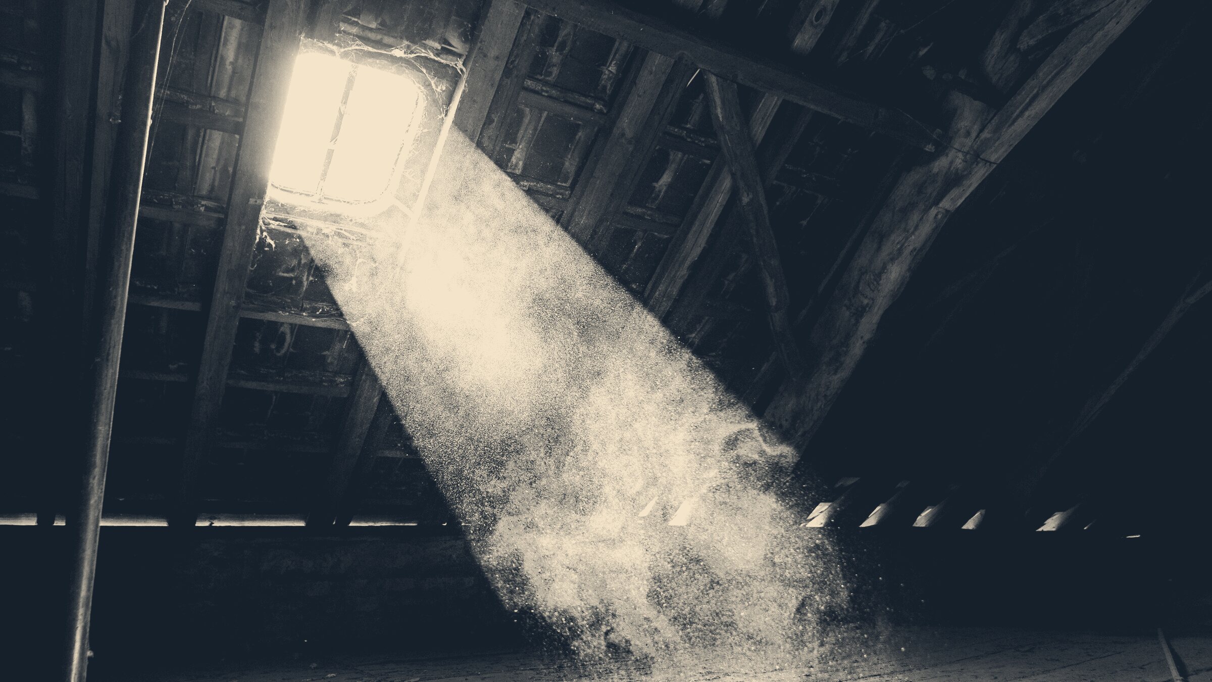 A shaft of sunlight through a broken attic skylight thick with swirling dust motes above dark timber floorboards