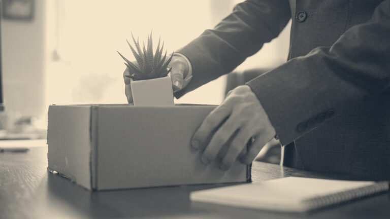 An employee in a grey suit packs a potted plant into a cardboard box on an office desk beside a notebook.