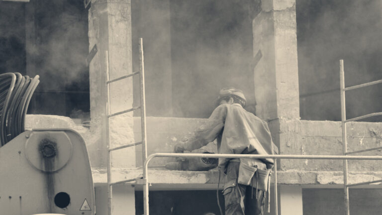 Worker in ear-defender helmet grinding a concrete beam amid dust on a construction site