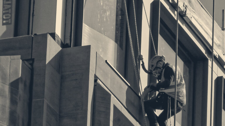 Rope-access worker in orange hi-vis and hard hat abseils down a City of London glass and stone office facade.
