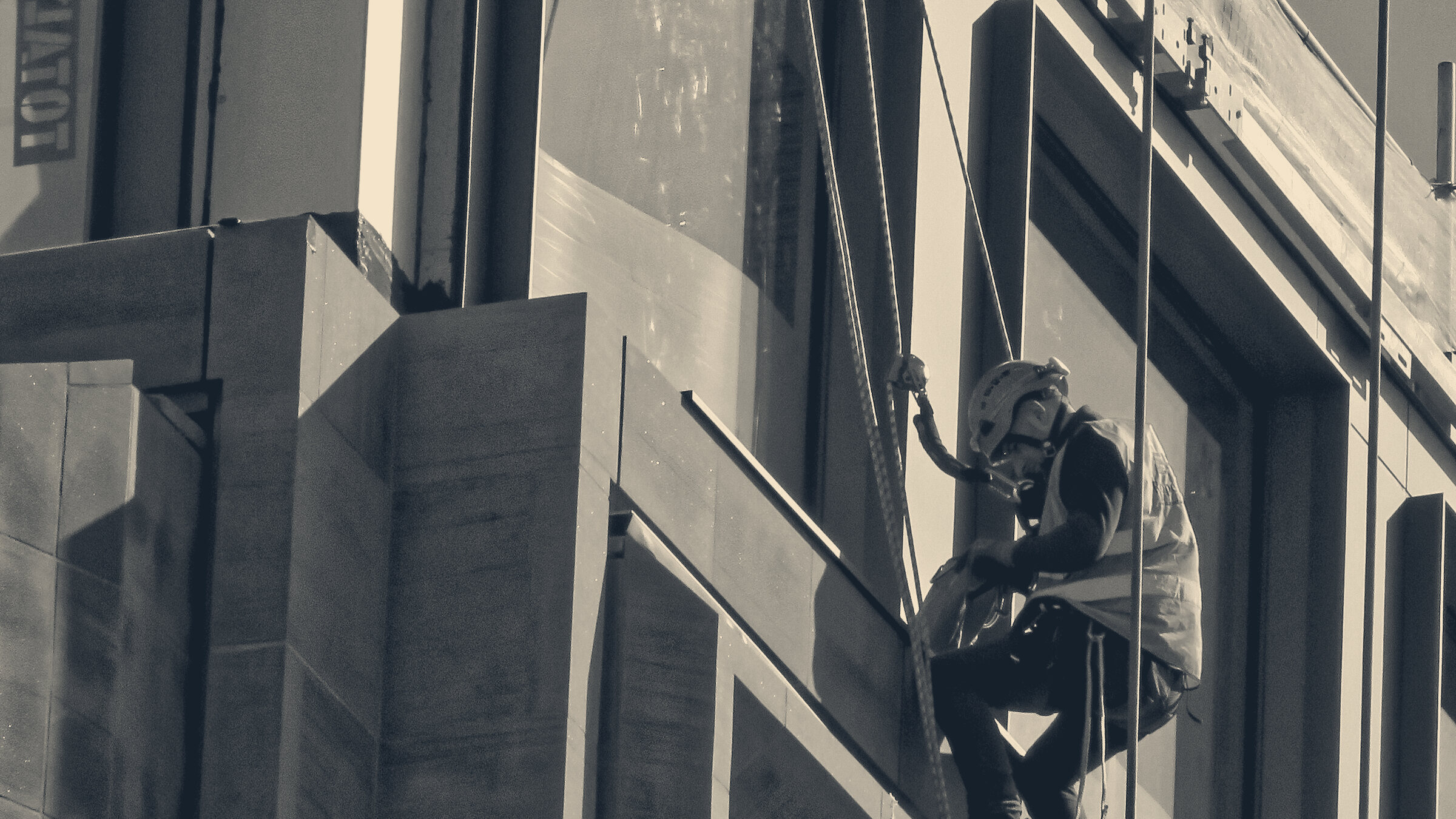 Rope-access worker in orange hi-vis and hard hat abseils down a City of London glass and stone office facade.