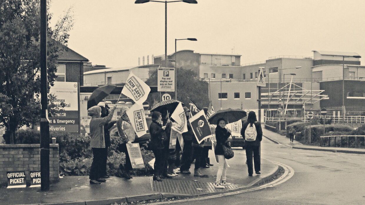 Midwives with umbrellas and RCM placards picketing outside West Middlesex Hospital entrance in the rain