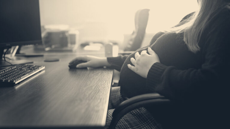 Pregnant woman in a black jumper at an office desk, hand on her bump, beside a monitor and keyboard