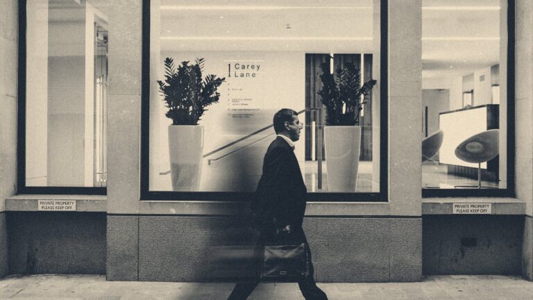 A man in a suit with a briefcase walks past the window of a City of London office building on Carey Lane.