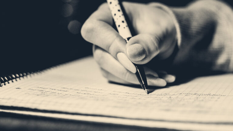 Hand gripping a ballpoint pen mid-sentence in a spiral-bound notebook, dark background