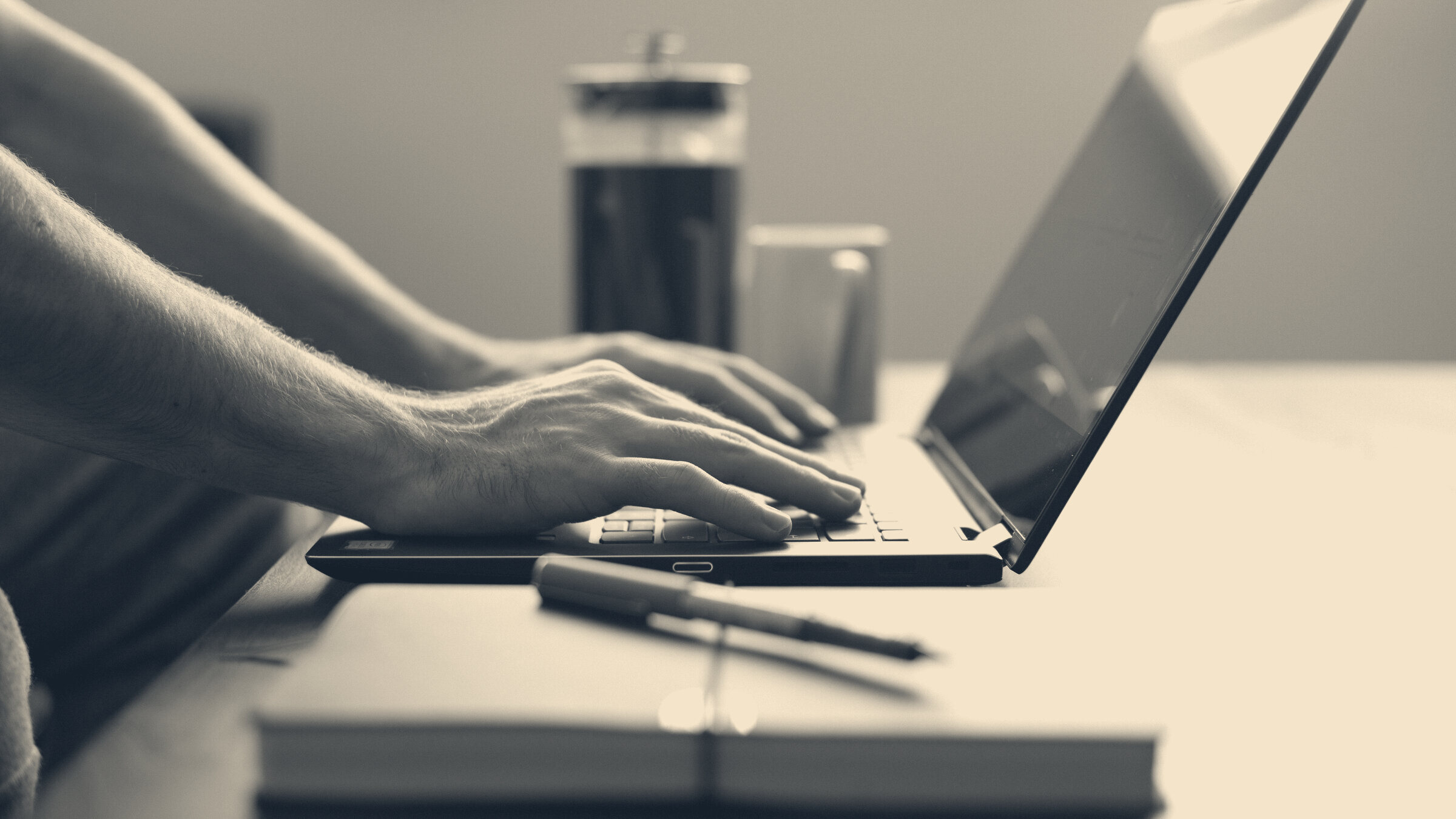 Hands typing on a laptop at a home desk, cafetiere and notebook in soft window light