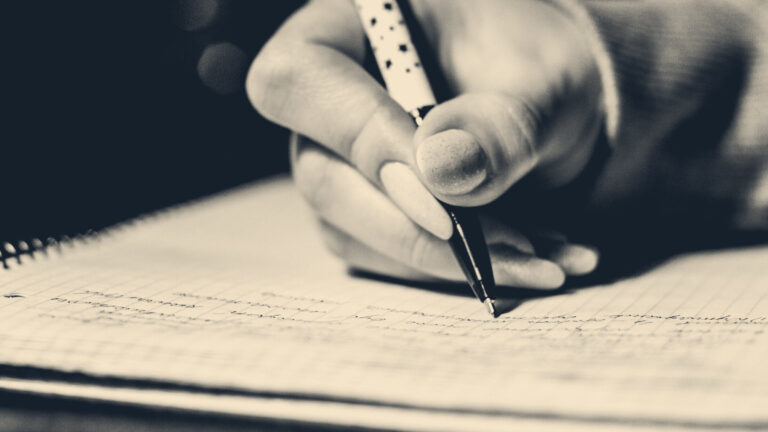 Close-up of a hand writing notes in a lined spiral notebook with a black-and-white pen