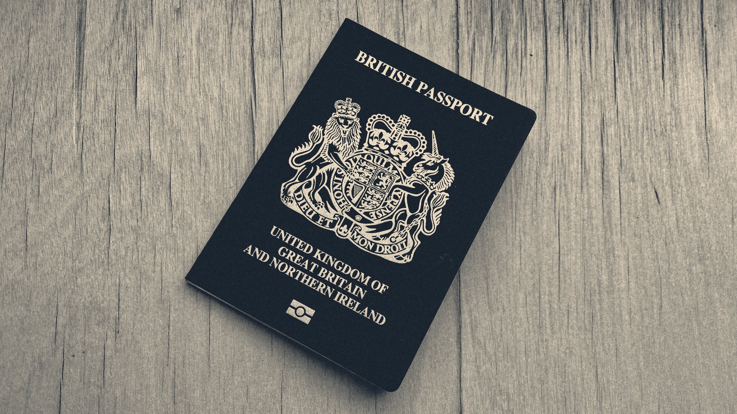 A navy British passport with gold crest lying tilted on a weathered pale-wood table.