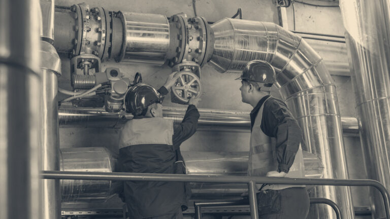 Two workers in hard hats and safety gear inspect and adjust an overhead valve on industrial plant piping.
