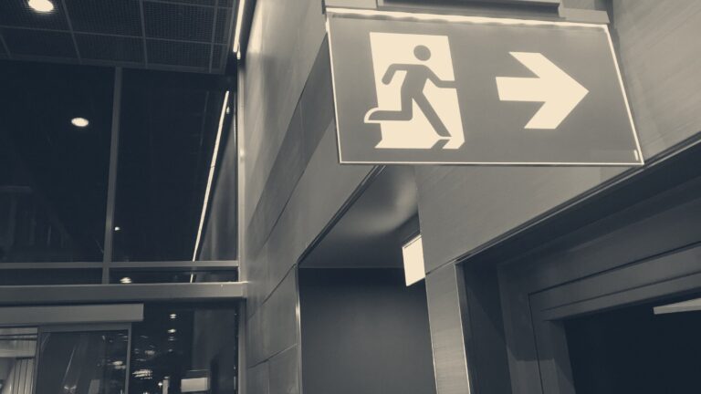 Illuminated green running-man emergency exit sign suspended from a ceiling in a dark corridor at night