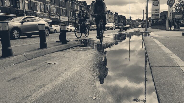 Two delivery riders cycle through a puddled bike lane on a Manchester street of red-brick shops.
