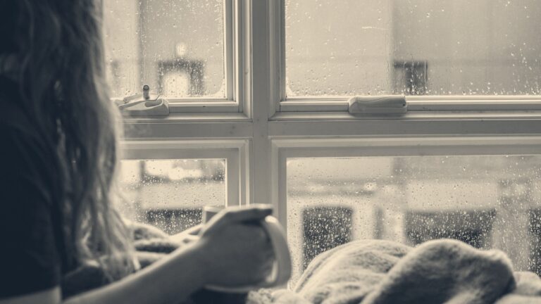Woman wrapped in a blanket holding a mug beside rain-streaked sash windows overlooking brick buildings