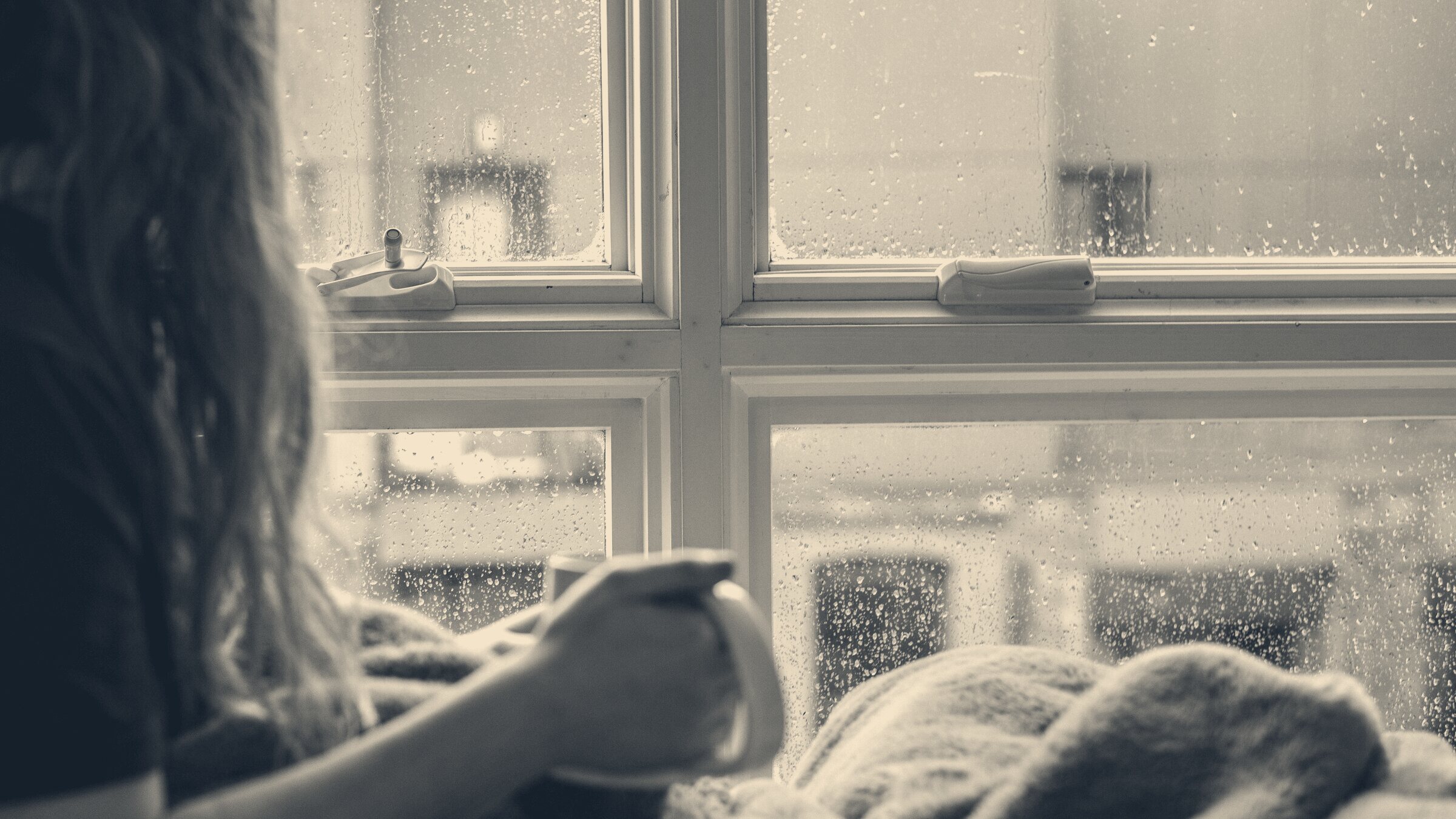 Woman wrapped in a blanket holding a mug beside rain-streaked sash windows overlooking brick buildings