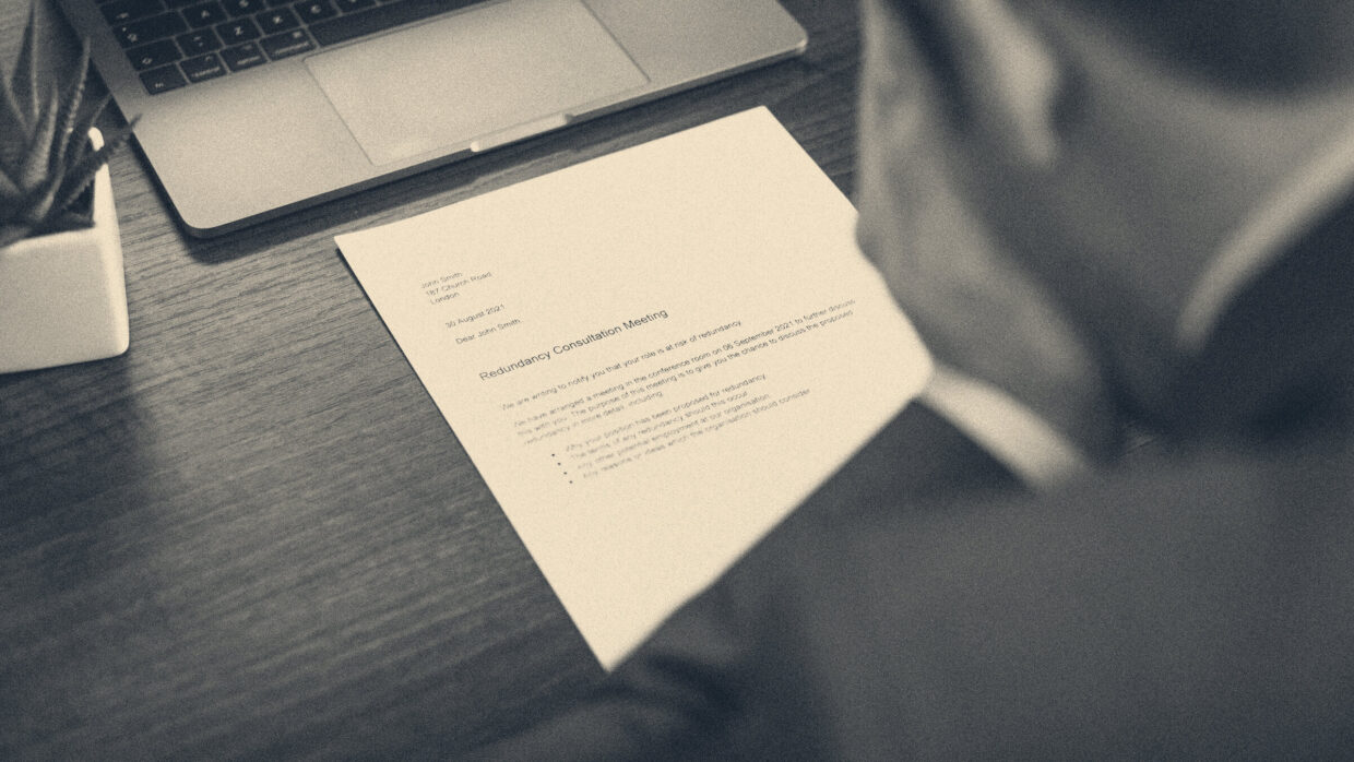 Over a man's shoulder, a formal employer's letter lies on a wooden desk beside a laptop.